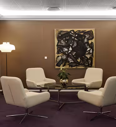 Ett modernt mötesrum med fyra krämfärgade stolar runt ett rektangulärt glasbord, flankerat av två vita golvlampor. En stor abstrakt målning hänger på den bruna väggen och en platt-TV är monterad på den motsatta vita väggen.