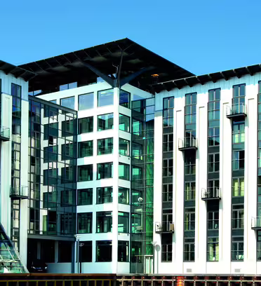 En modern flervåningsbyggnad med stora glasfönster och balkonger, med en kombination av metall- och glasarkitektur. Strukturen inkluderar ett centralt glasatrium och har en ljus, klarblå himmel i bakgrunden.