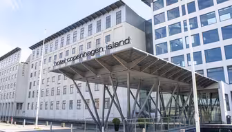 Modern byggnad exteriör på Hotel Copenhagen Island, med stora fönster och en unik entrétak som stöds av kantiga metallbjälkar. Hotellets namn är väl synligt ovanför entrén.