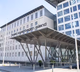 Modern byggnad exteriör på Hotel Copenhagen Island, med stora fönster och en unik entrétak som stöds av kantiga metallbjälkar. Hotellets namn är väl synligt ovanför entrén.
