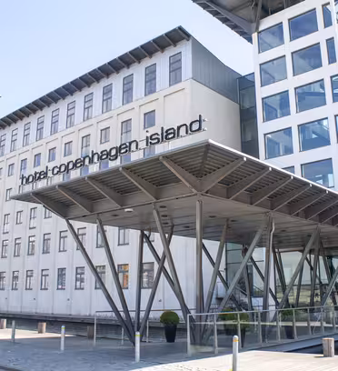 Modern byggnad exteriör på Hotel Copenhagen Island, med stora fönster och en unik entrétak som stöds av kantiga metallbjälkar. Hotellets namn är väl synligt ovanför entrén.