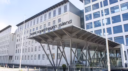 Modern byggnad exteriör på Hotel Copenhagen Island, med stora fönster och en unik entrétak som stöds av kantiga metallbjälkar. Hotellets namn är väl synligt ovanför entrén.