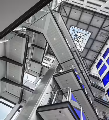 Interiörvy av en modern byggnad med flera trappor och balkonger i ett atrium. Designen har glas- och metallelement, med ett takfönster upptill. Stora blå fönster ger kontrast och skapar en öppen och futuristisk atmosfär.