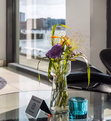 En vas med färgglada blommor står på ett glasbord bredvid en liten blå ljushållare och ett kort. I bakgrunden är moderna svarta stolar placerade nära stora fönster med utsikt över en stadsbild och vatten.