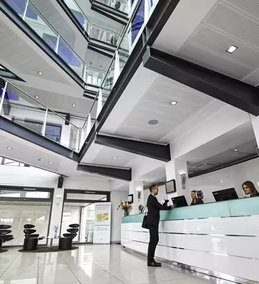 En modern kontorslobby med en vit reception. Två sittande receptionister hjälper en stående person. Lokalen har högt i tak, ett glasatrium, flera våningar med räcken och modern inredning, inklusive abstrakt konst och eleganta möbler.