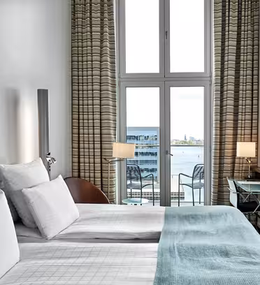 Modernt hotellrum med två sängar klädda i vita sängkläder och en kricka. Ett stort fönster erbjuder utsikt över vattnet. Rummet har en väggmonterad TV, randiga gardiner och ett litet skrivbord med lampa.