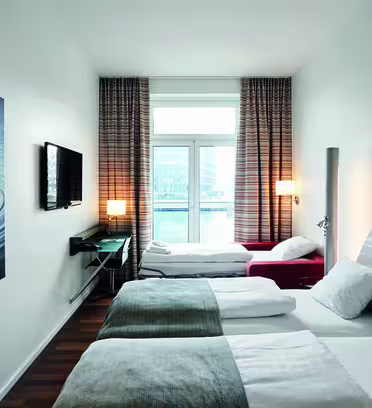 Modernt hotellrum med två sängar, med grå och vita detaljer. På väggen hänger ett stort abstrakt konstverk som föreställer vattenkrusningar. En platt-TV finns i närheten. Ett fönster med gardiner från golv till tak har utsikt över stadsbilden.