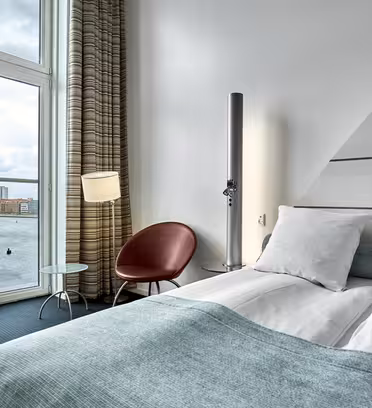 Modernt hotellrum med dubbelsäng, ljusblå sängkläder, cylindrisk golvlampa och rund röd stol. Ett litet glasbord står vid fönstret, som erbjuder utsikt över en molnig himmel över en stadsbild och vatten.