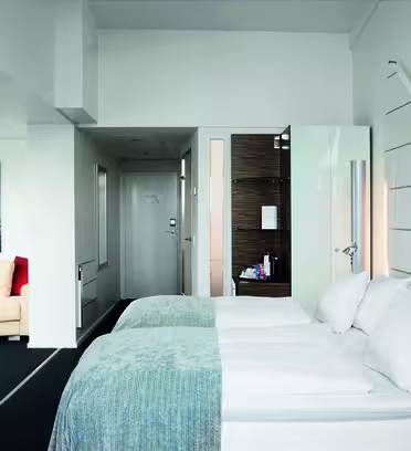 Hotellrum med två prydligt bäddade enkelsängar klädda i ljusblåa plagg och vita kuddar. En väggmonterad TV finns till vänster och en liten sittgrupp med en beige soffa syns. Ett modernt badrum syns delvis nära entrén.