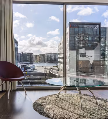 Modernt vardagsrum med ett stort fönster med utsikt över stadsarkitektur och en kanal. Rummet har ett runt soffbord i glas, en röd stol, en grå soffa och en texturerad matta. Starkt dagsljus lyser upp utrymmet och framhäver den eleganta inredningen.