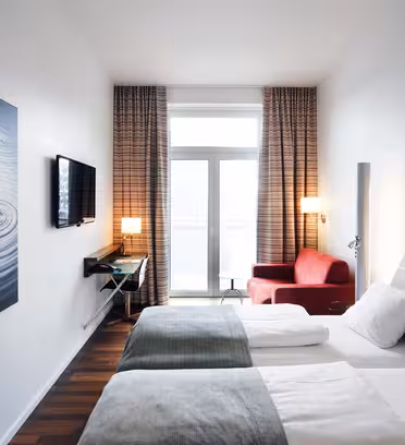 Ett modernt hotellrum med två snyggt bäddade sängar, en väggmonterad TV och ett stort fönster med gardiner. En röd fåtölj och ett inramat konstverk som visar vattenkrusningar bidrar till inredningen. Rummet har mörka trägolv.