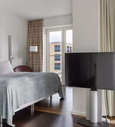 Modernt sovrum med en stor säng, en golvlampa och en TV på stativ. Rummet har fönster från golv till tak med utsikt över vattnet, inklusive byggnader och en molnig himmel. Gardiner hänger vid sidan av fönstret.