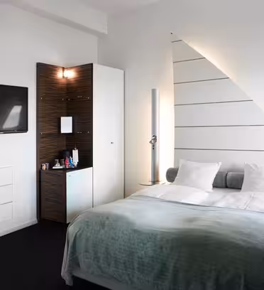 Ett modernt hotellrum har en snyggt bäddad dubbelsäng med vita sängkläder och en kricka. En platt-TV är monterad på väggen. Rummet har elegant inredning, inklusive en triangulär väggdesign och ett litet skåp fyllt med förfriskningar.