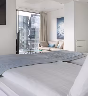 Modernt hotellrum med en snyggt bäddad säng och en ljusblå filt. Det finns en soffa med kuddar nära ett stort fönster som erbjuder utsikt över stadsbilden och en byggnad märkt "Handels". Rummet har minimalistisk inredning med en väggmonterad TV.