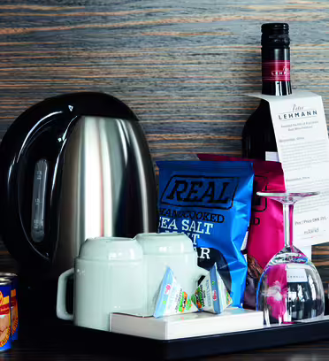 En bricka för förfriskningar på hotellrummet med en vattenkokare i rostfritt stål, konserverade persikor, tekoppar, tepåsar, två glas, en flaska vin och ett paket havssaltchips, mot en tränäringsbakgrund.
