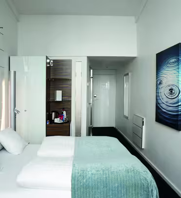 Ett modernt hotellrum med vita väggar och minimalistisk inredning. Den har en snyggt bäddad säng med ett ljusblått plagg, en väggmonterad TV och konstverk av koncentriska cirklar. En liten öppen garderob och en dörröppning syns i bakgrunden.