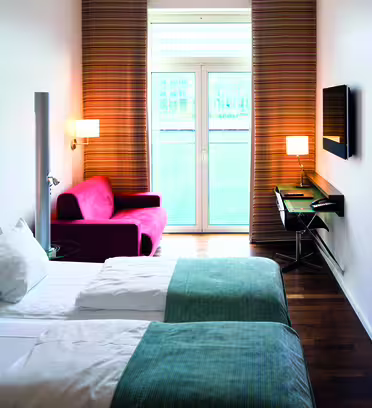Ett modernt hotellrum med två enkelsängar, en röd fåtölj och en platt-TV på väggen. Det finns ett skrivbord med en stol bredvid TV:n. Blåvita sängkläder, randiga gardiner och en stor abstrakt målning syns.