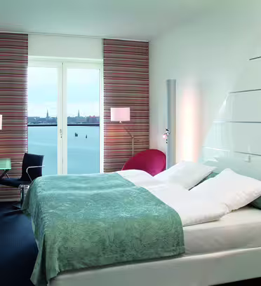 Ett modernt hotellrum med en stor säng täckt av en ljusblå och vit filt. Det finns en röd stol, ett skrivbord och en stol vid en väggmonterad TV. Fönstret erbjuder utsikt över en vattenmassa och byggnader i fjärran.