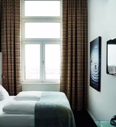 Ett modernt hotellrum med en snyggt bäddad säng, en väggmonterad platt-TV och ett stort fönster med randiga gardiner. Det finns ett horisontellt konstverk på väggen och ett bord med en telefon i förgrunden.