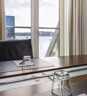 Ett modernt konferensrum med träbord, läderstol, anteckningsblock, penna och inverterat glas. Ett stort fönster visar en vy av en vattenmassa och byggnader utanför. Gräddgardiner ramar in fönstret och en klädkrok sitter på väggen.