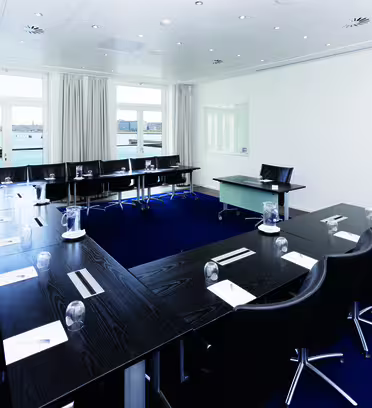 Ett modernt konferensrum med en U-formad svart bordsuppsättning, omgiven av svarta stolar. Anteckningsblock och glas ställs på bordet. Stora fönster ger utsikt över en vattenmassa och byggnader. Rummet är väl upplyst och har vita väggar.
