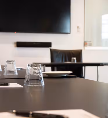 Ett konferensrum med stor skärm på väggen. Ett bord är dukat med upp-och-nervända glasögon, pennor och anteckningsblock. Svarta stolar står uppställda runt bordet. Rummet har stark belysning och minimalistisk design.