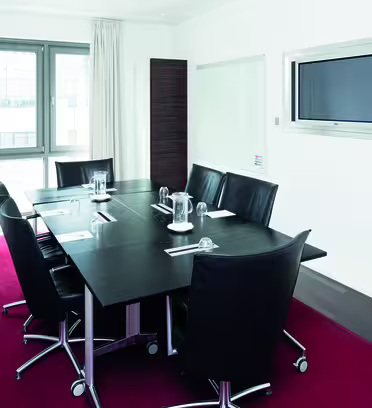 Ett modernt konferensrum med ett rektangulärt svart bord, sex svarta läderstolar och en inbyggd platt-TV på väggen. Rummet har vita väggar, stora fönster med vita gardiner och en röd matta.
