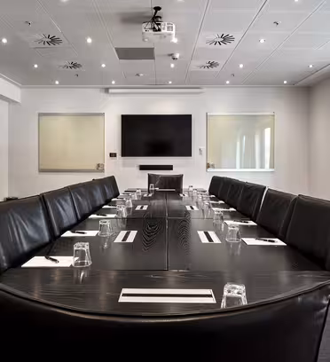 Ett modernt konferensrum med ett stort svart rektangulärt bord omgivet av svarta läderstolar. Bordet är dukat med anteckningsblock och glas. En platt-TV är monterad på väggen, med två whiteboardtavlor på vardera sidan. Ljus belysning och fönster.