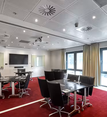 Modernt konferensrum med röd matta, med svarta bord och stolar. Stora fönster med gardiner kantar ena sidan, och en projektor och duk finns på framsidan. Taklampor och luftventiler är synliga, vilket skapar ett väl upplyst och organiserat utrymme.