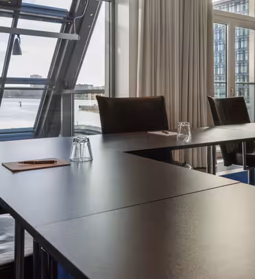 Ett modernt konferensrum med ett U-format bordsarrangemang. Varje bord har ett anteckningsblock och ett upp och nedvänt glas. Stora fönster ger utsikt över vattnet och stadsbilden. Naturligt ljus fyller rummet genom skira vita gardiner.