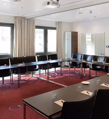 Ett modernt konferensrum med U-formade svarta bord och stolar på en röd matta. Varje inställning har ett anteckningsblock och glas. Stora fönster med gardiner, en väggmonterad TV, en projektor och vita väggar fullbordar installationen.