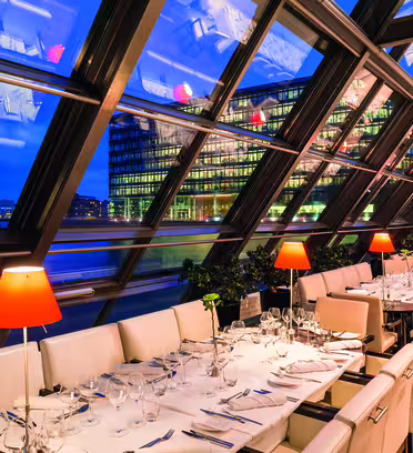 En modern restaurang med en glasvägg med utsikt över en stadsbild vid vattnet. Interiören har vita vadderade stolar, snyggt dukade bord med vinglas och röda bordslampor. Skylinen reflekteras på glaset, vilket ger en dynamisk stadsvy.