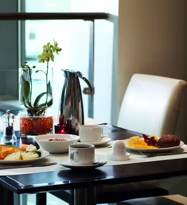 Ett väldukat bord för frukost med en vit duk, med en kopp kaffe, skivad frukt, bröd, äggröra och ett mjukkokt ägg. Det finns också kryddor, en krukväxt och en karaff på bordet.