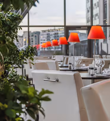 Modern restauranginteriör med vita stolar och träbord med glas och bestick. Röda lampskärmar kantar borden och skapar en varm atmosfär. Stora fönster erbjuder utsikt över stadsbilden och en krukväxt lägger till grönska i förgrunden.