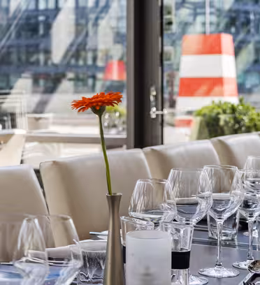 Ett elegant restaurangbord dukat med tomma vinglas och vita servetter på en terrass. Två orangea blommor i vaser ger en nyans av färg. I bakgrunden avslöjar höga fönster en geometrisk byggnad och rödrandig struktur utanför.