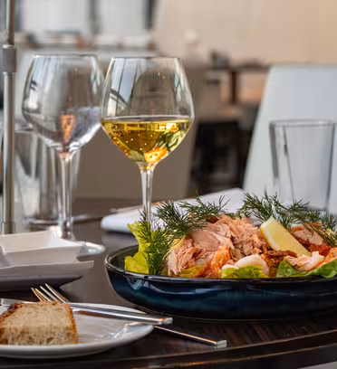 Ett bord dukas med två glas vitt vin, en skål sallad toppad med lax och citronklyftor, och brödskivor på tallrikar. Inramningen är en modern, elegant restaurang med mjuk belysning.