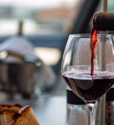 Ett glas rött vin hälls upp från en flaska i en elegant miljö. I förgrunden står brödskivor. I bakgrunden vilar en flaska i en ishink på ett bord. Scenen antyder en sofistikerad matupplevelse.