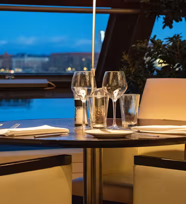 Ett mysigt restaurangbord för fyra, med vinglas, bestick och servetter. Mjuk belysning skapar en varm atmosfär. Ett stort fönster i bakgrunden erbjuder utsikt över en kvällsstadsbild och blå himmel.