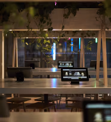 Modern kaféinteriör med bord med interaktiva skärmar och trästolar. Svävande växter ger grönska till atmosfären. Stora fönster i bakgrunden avslöjar en antydan av gatubelysning och en neonskylt utanför.