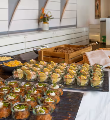 Ett buffébord dukas med olika aptitretare, inklusive små glasburkar fyllda med krämiga rätter, garnerade med örter. Tallrikar med bröd, citronklyftor och sallader visas också. En vas med blommor pryder bakgrunden.