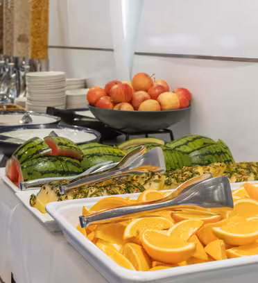 En frukostbuffé med brickor med skivade apelsiner, ananas och vattenmelon. Bakom frukten finns automater med flingor, en bunt tallrikar, en glasskål med hårdkokta ägg och en skål med äpplen på en vit duk.