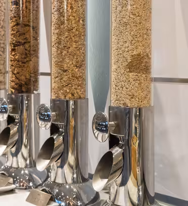 Rad av spannmålsautomater fyllda med olika spannmål, inklusive granola och cornflakes. Var och en har en metallbas och pip för enkel dispensering, med små etiketter framtill. De står uppställda på en bänkskiva mot en kaklad vägg.
