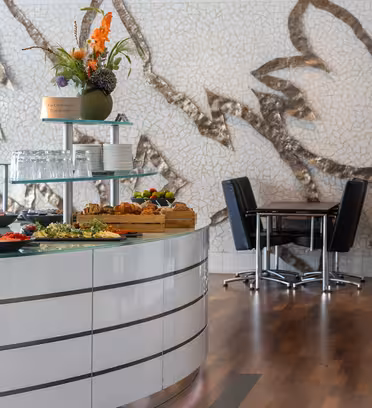 En modern matplats med en vit böjd bänkskiva med diverse rätter som bakverk och frukt. Bakom den en mosaikväggkonst och en liten vas med blommor. Till höger ett bord med svarta stolar. Golvet är trä, vilket ger värme till utrymmet.