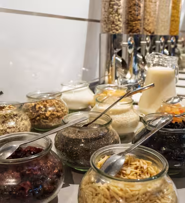Ett frukostbuffébord med olika glasburkar fyllda med ingredienser som nötter, torkad frukt och frön. I bakgrunden finns automater med flingor, en kanna mjölk och staplade tallrikar med redskap.
