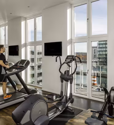 En person tränar på ett löpband i ett modernt gym med stora fönster som visar en stadsbild och vattenvy. Olika gymutrustningar är synliga, med naturligt ljus som lyser upp utrymmet.