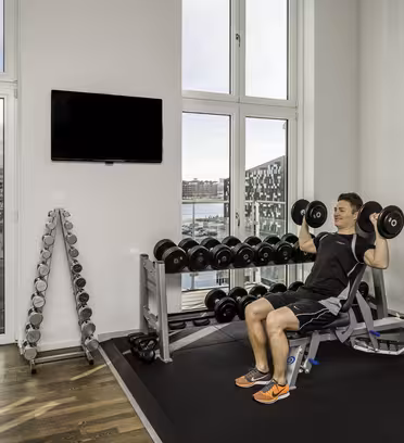 En person i atletisk kläder sitter på en bänk i ett gym och lyfter hantlar ovanför sina axlar. Rummet har stora fönster med utsikt över stadsbild och vatten. En uppsättning hantlar syns på ställ längs väggen.