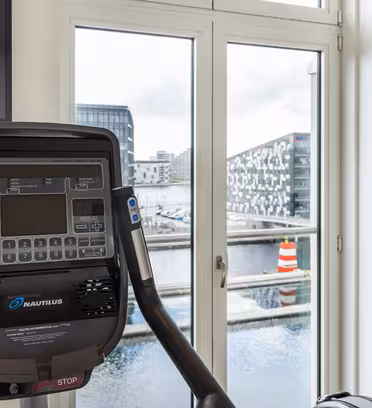 En stationär motionscykel med elektronisk display är placerad i ett modernt rum. Stora fönster visar en utomhusvy av en vattenmassa och moderna byggnader i bakgrunden.