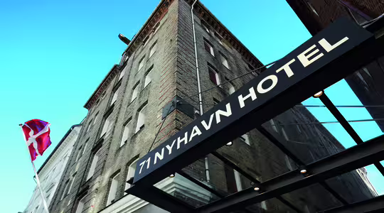 Bilden visar en hög tegelbyggnad med en skylt med texten "71 Nyhavn Hotel" på en baldakin. En dansk flagga vajar på en stång till vänster mot en klarblå himmel.