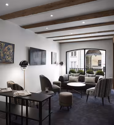 Modern lounge med beiga stolar och små bord på en mörk matta. Exponerade träbjälkar och vita väggar har abstrakt konst. Stora fönster erbjuder utsikt över gatan, vilket skapar en mysig och inbjudande atmosfär.