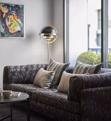 Mysigt vardagsrum med en mörkmönstrad soffa prydd med kuddar, ett runt soffbord med en vit kopp och skål, modern golvlampa, abstrakt väggkonst och ett stort fönster som visar en balkong med krukväxter.